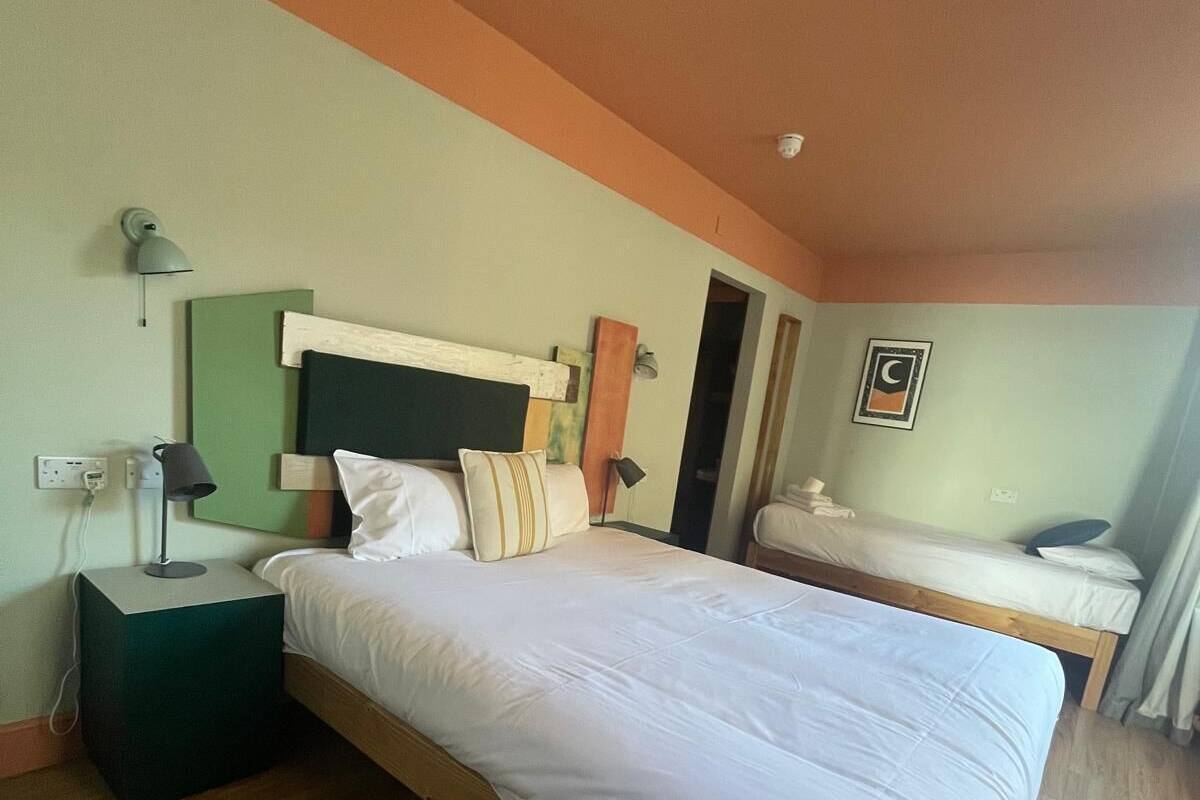 A hotel room with a large bed, a desk, and a chair. There is a nightstand with a lamp, a wall-mounted shelf, and a framed picture.
