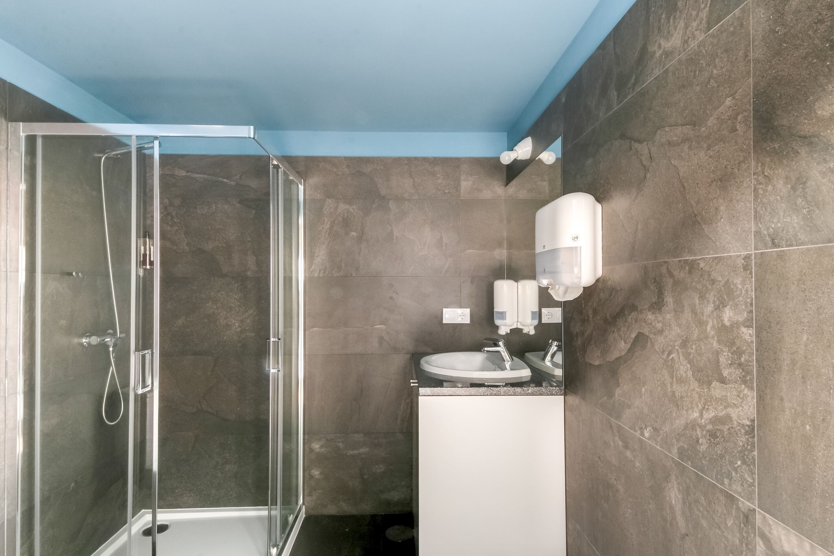 A bathroom with a glass-enclosed shower, a sink, and a soap dispenser mounted on the wall.