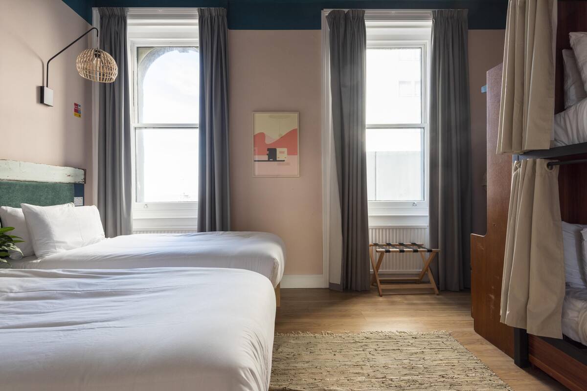 A bedroom with two beds, a wooden floor, a woven rug, a wall-mounted lamp, and a window with curtains.
