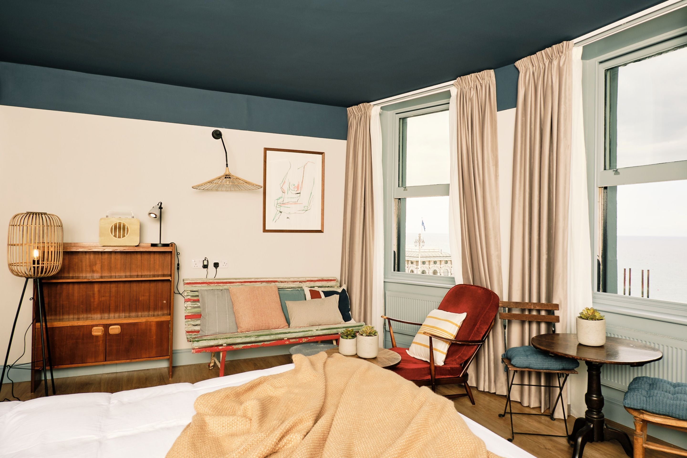 A bedroom with a bed, a wooden wardrobe, a red armchair, a small table with a plant, and a window with curtains.