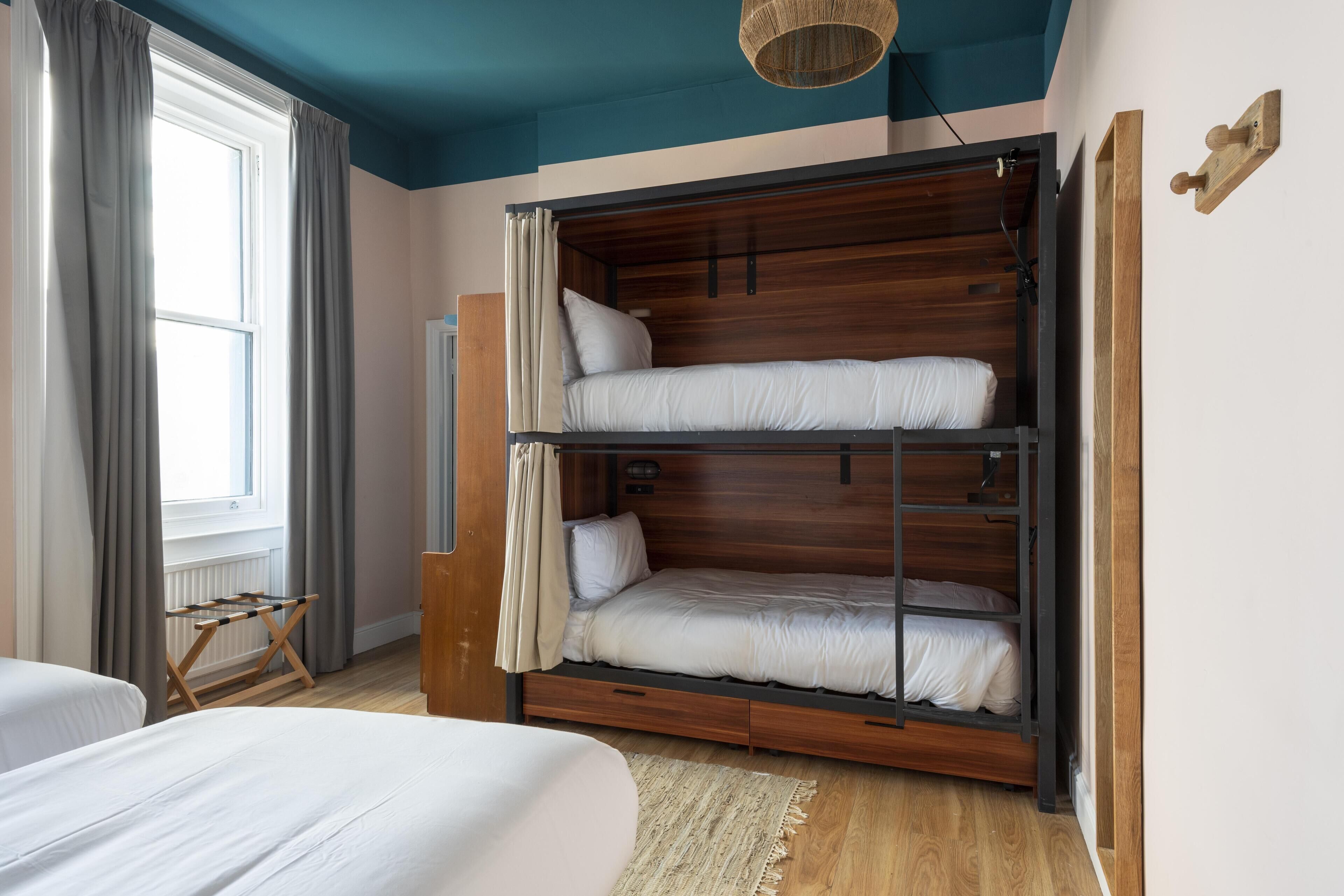 A room with a bunk bed, a window with curtains, a wooden floor, and a hanging light fixture.