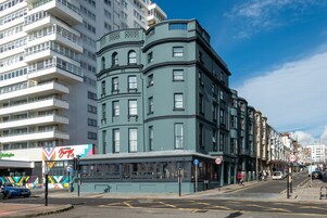 A multi-story building with a corner entrance, a bus stop, and a street with cars and pedestrians.