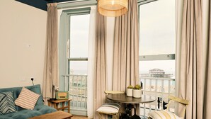 Een knus interieur met een bed, een houten tafel met planten, een blauwe bank en uitzicht op het strand door grote ramen.