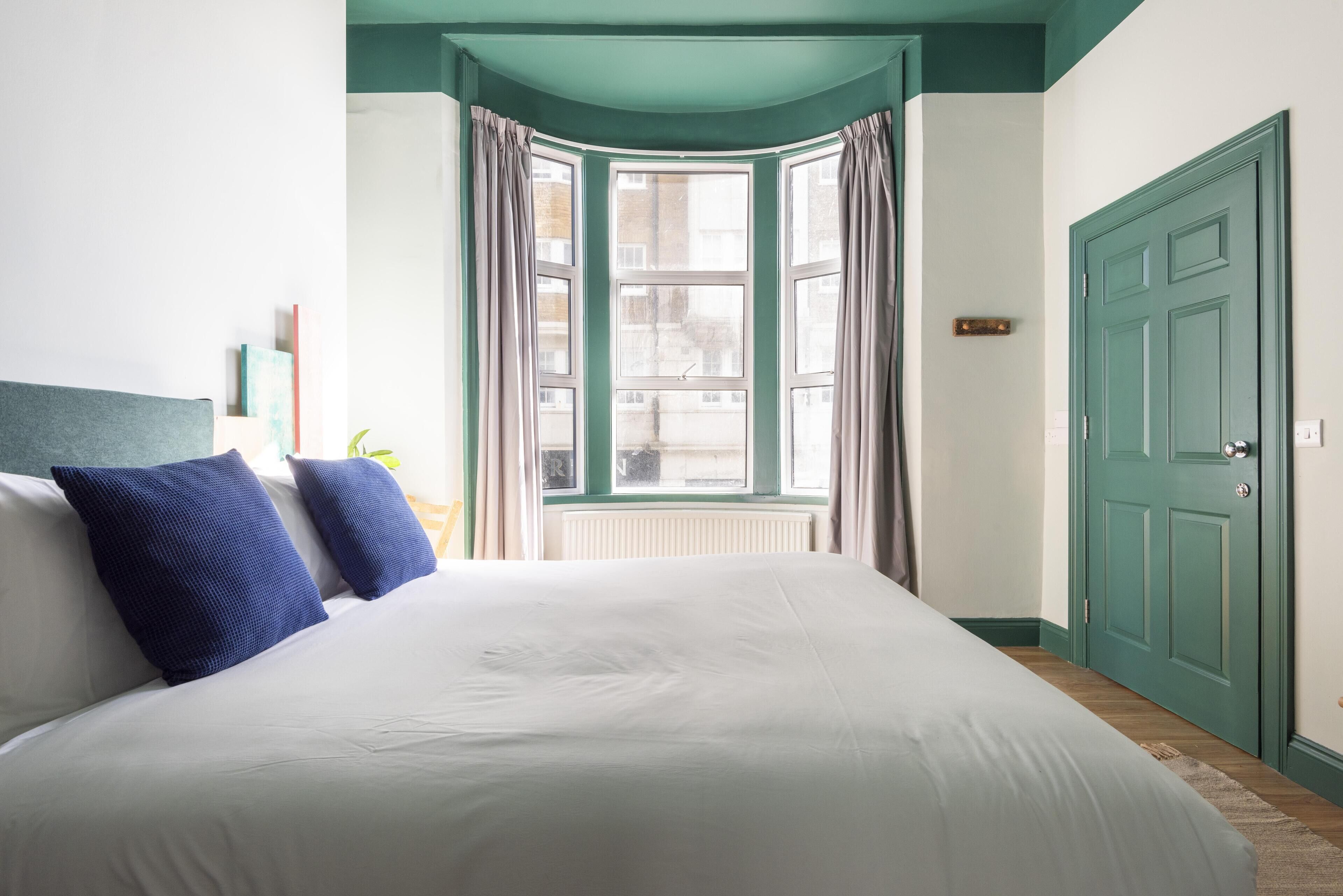 Une chambre avec un grand lit, une fenêtre en saillie avec des rideaux, une porte verte et un radiateur.