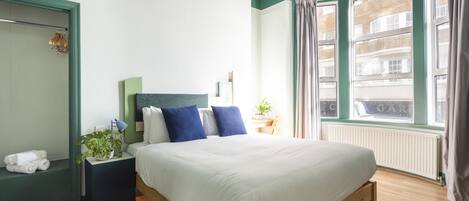 A neatly arranged bedroom with a large bed, a bedside table with a lamp, a small plant, and a view of the city through a window.