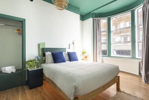 A neatly arranged bedroom with a large bed, a bedside table with a lamp, a small plant, and a view of the city through a window.