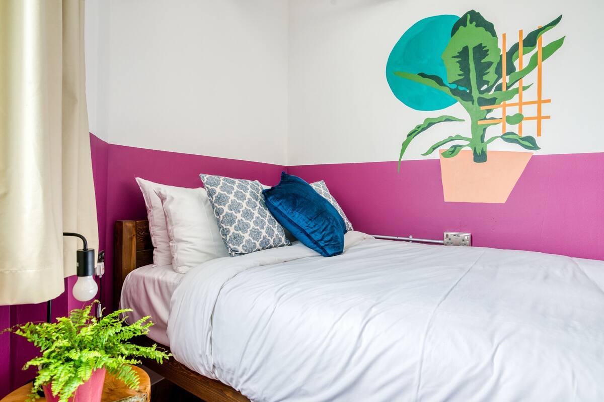 A bedroom with a bed, pillows, a wall art of a plant, a bedside table with a lamp, and a potted plant.