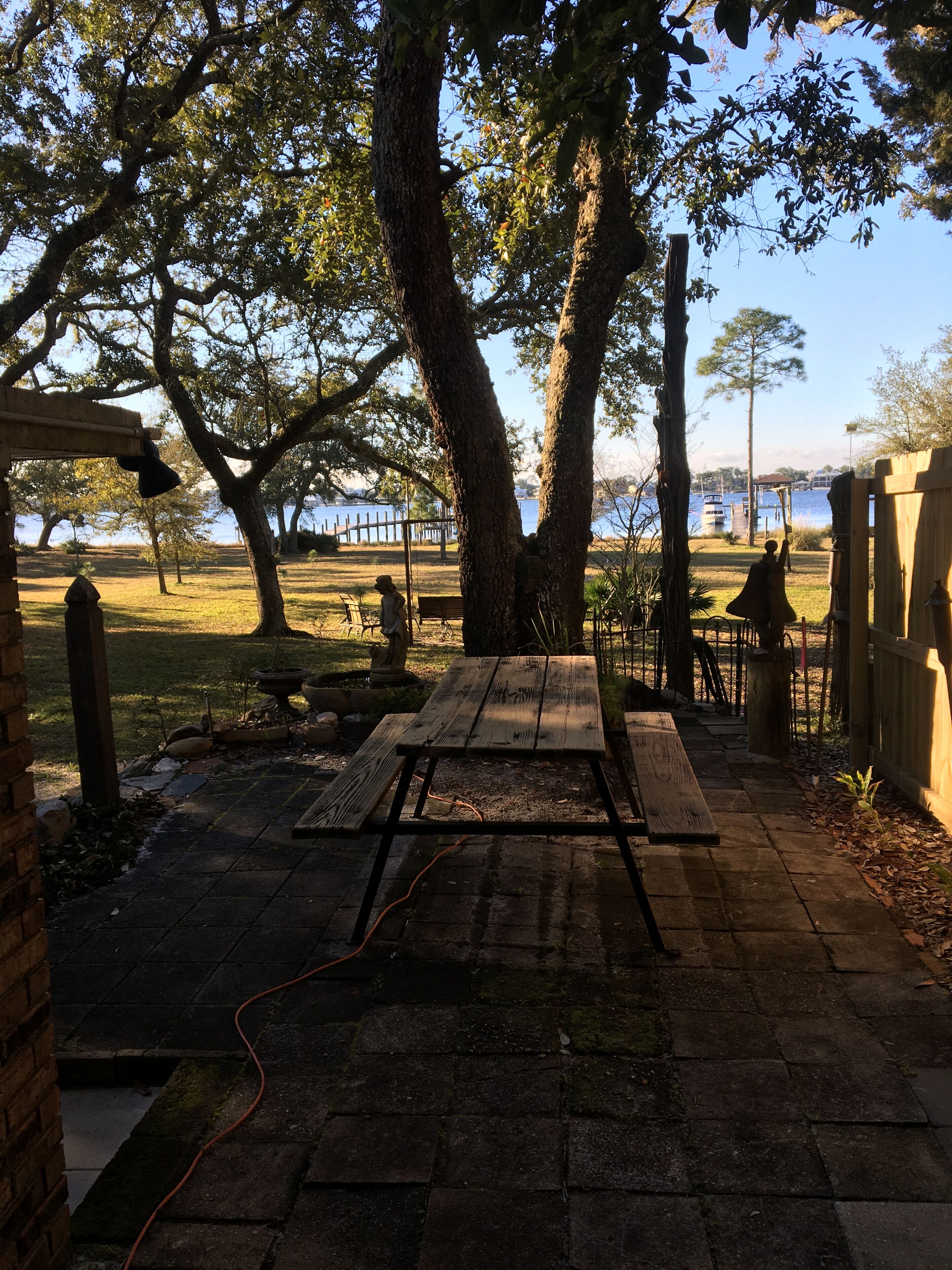 A wooden picnic table under a tree, a brick pathway, and a view of a body of water with boats.