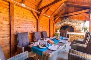 Een buitenterras met een lange tafel gedekt voor een maaltijd, omringd door rieten stoelen en een stenen open haard.