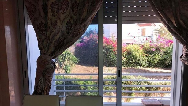 A balcony with a table and chairs, a view of a garden with pink flowers, and a building in the background. - Appart Residence Al Menzah (Agadir)