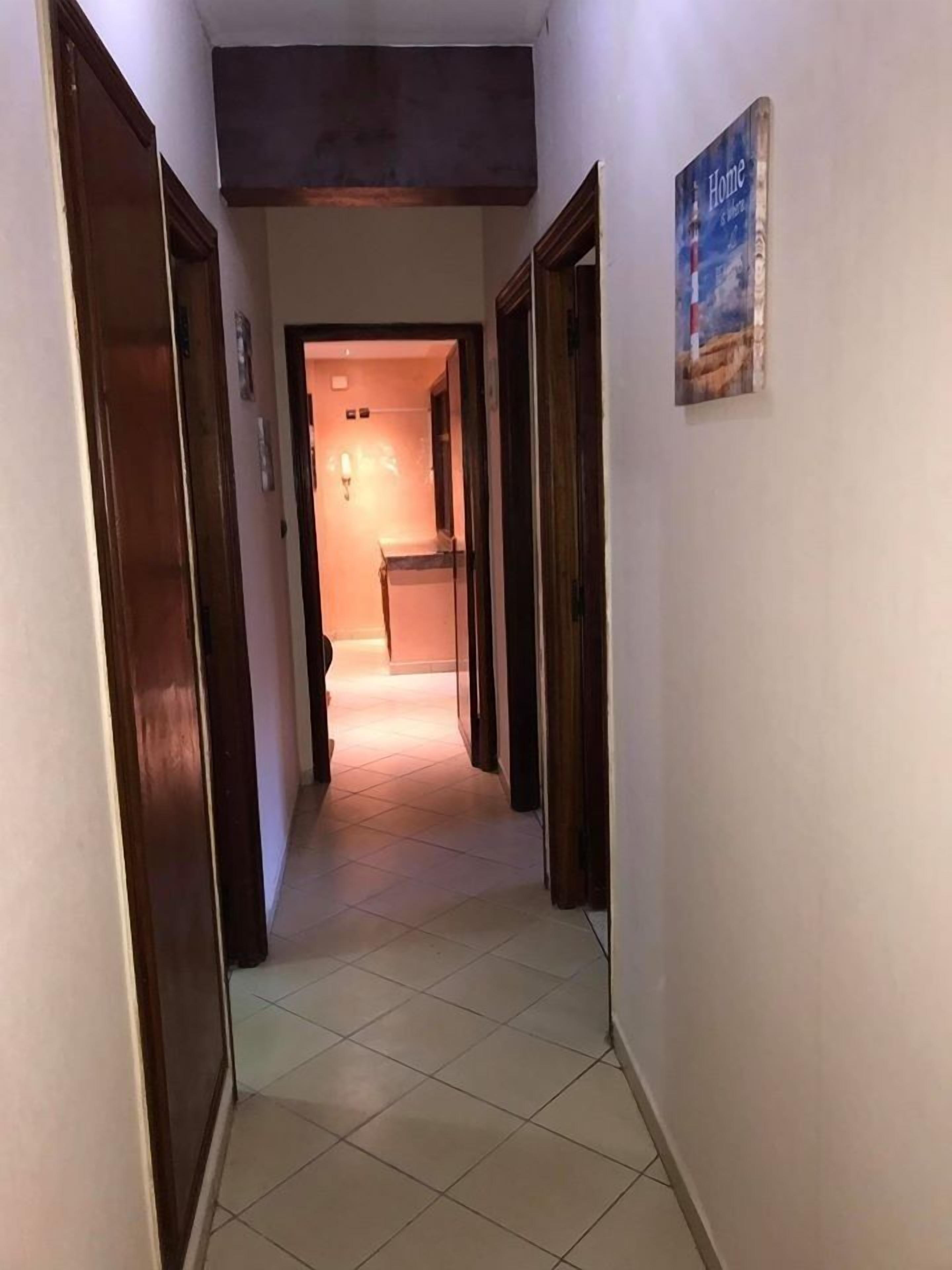 A hallway with tiled flooring and wooden doors, leading to a room with a lit interior.