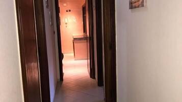 A hallway with tiled flooring and wooden doors, leading to a room with a lit interior.