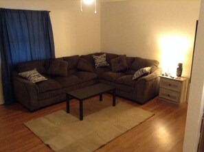 A living room with a sectional sofa, a coffee table, a side table with a lamp, and a blue curtain. - Convenient 1-Bedroom Apartment Next to Fort Sill (Lawton)