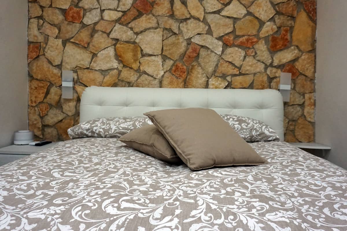 A bedroom with a stone wall, a tufted headboard, patterned bedding, and a bedside table with a lamp.