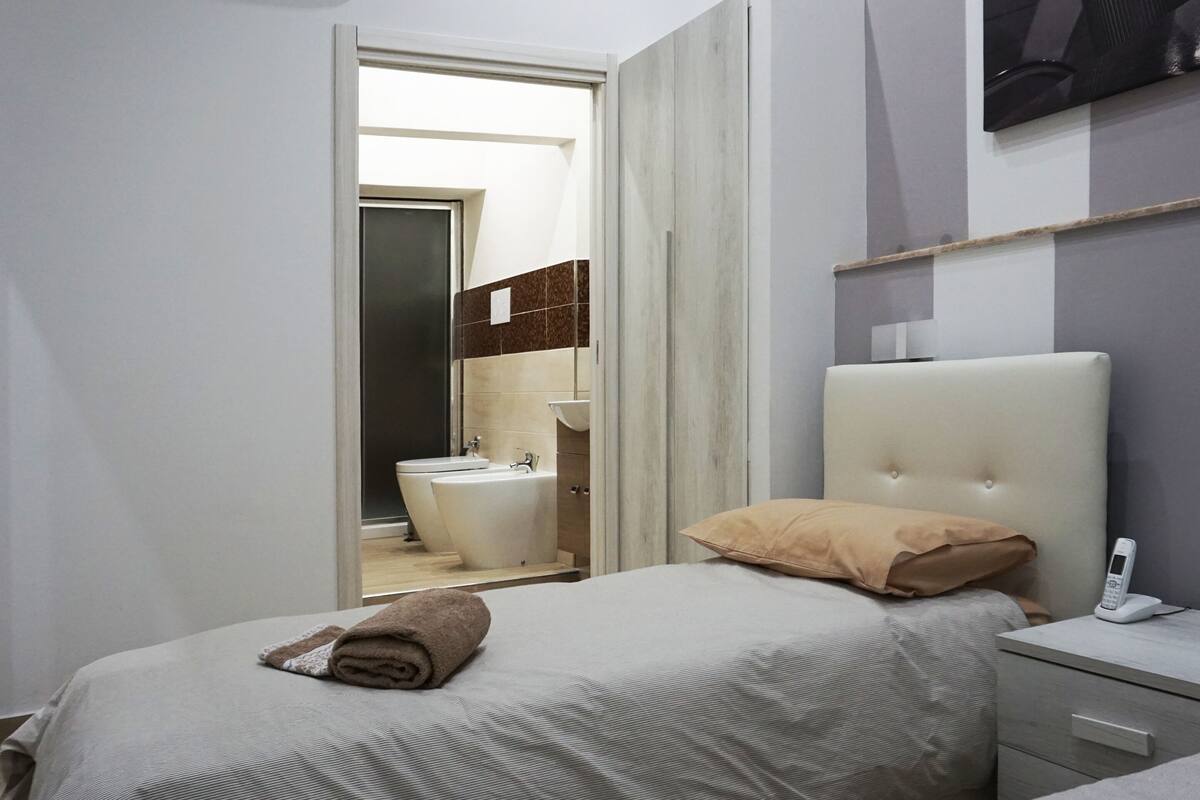 A bedroom with a bed, a bedside table with a phone, and a view of a bathroom with a sink and toilet.