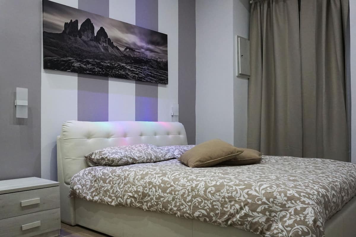 A bedroom with a bed, a nightstand, a drawer unit, a painting on the wall, and a window with curtains.