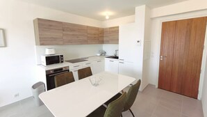 Una cocina moderna con armarios blancos, encimera blanca, microondas, cafetera y un lavabo. Hay una mesa de comedor con sillas y una puerta a la derecha.