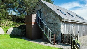 A stone building with a wooden staircase leading to a door, surrounded by a fenced area and large rocks. - Dartmoor Barn on North Hessary Tor (Yelverton)