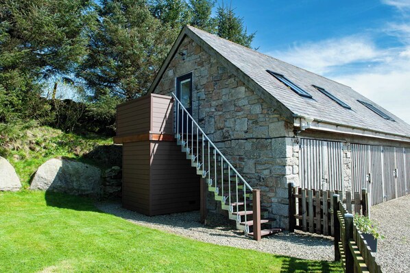 A stone building with a wooden staircase leading to a door, surrounded by a fenced area and large rocks.
