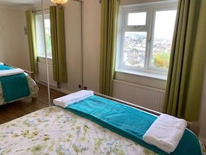 Uma cama de solteiro com colcha verde-água e toalhas brancas, uma janela com vista para uma área residencial e cortinas verdes.
