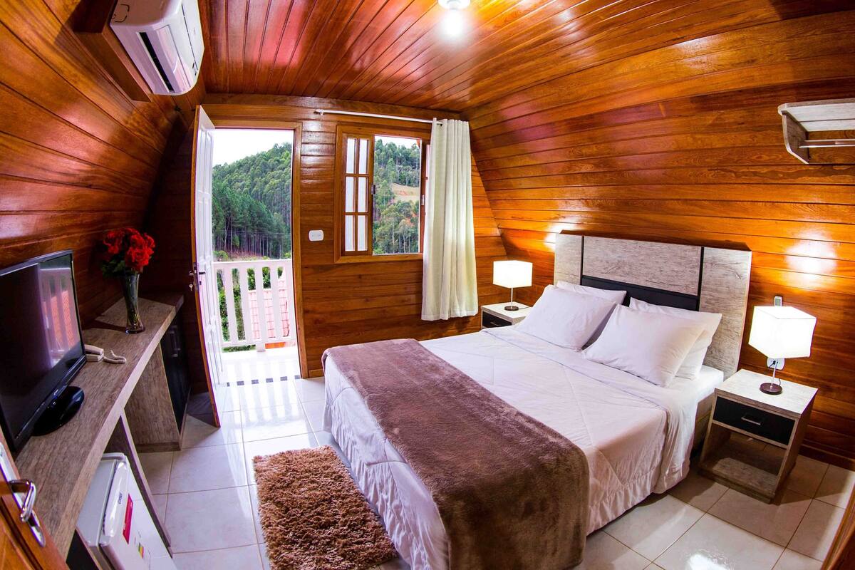 Een kamer met houten lambrisering, een bed, bedlampen, een televisie en uitzicht op een balkon met een hekwerk.