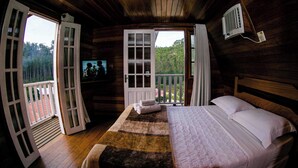 A wooden cabin bedroom with a bed, a TV, and a balcony with a view of the forest. - China Park Eco Resort (Domingos Martins)