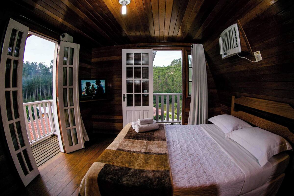 Een houten slaapkamer met een bed, een televisie en een balkon met uitzicht op het bos.