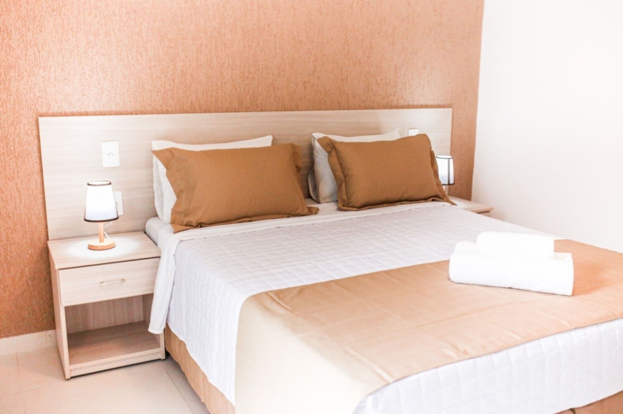Un letto rifatto con un copriletto bianco e due cuscini marroni, un comodino con una lampada e un interruttore luce a parete.
