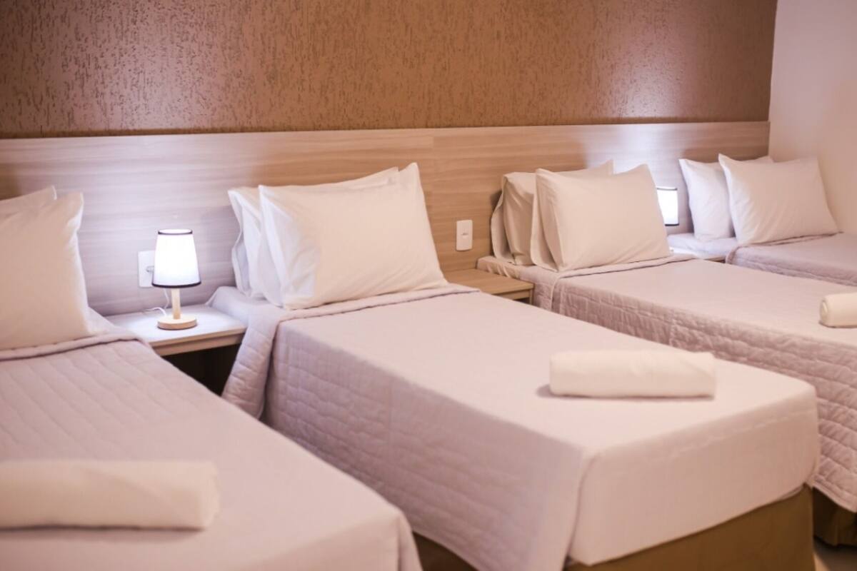 Een hotelkamer met drie eenpersoonsbedden, elk met een witte deken en kussen, een nachtkastje met een lamp, en een houten hoofdbord.