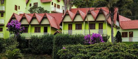A row of green buildings with red roofs and white balconies, surrounded by lush greenery and purple flowers.