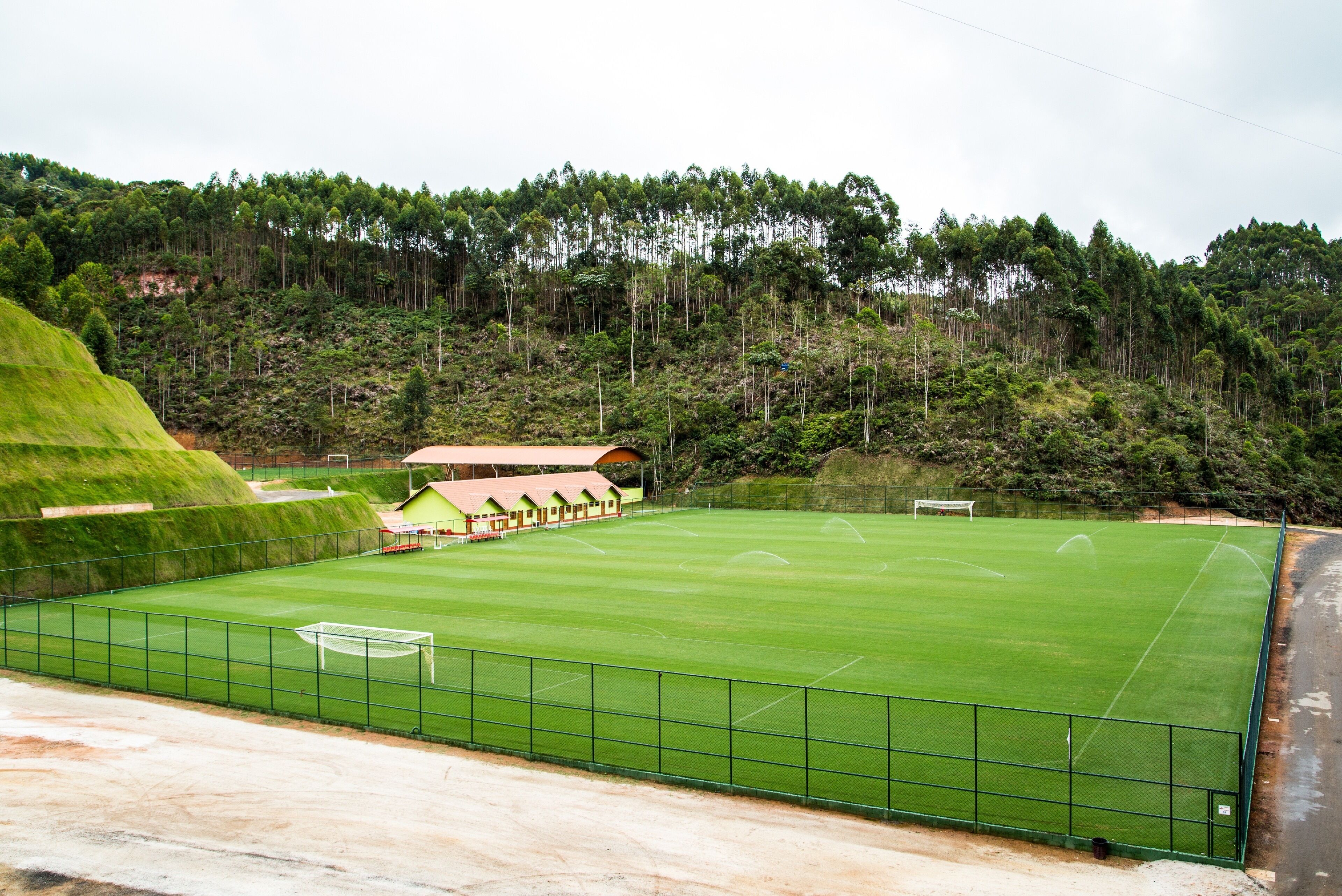 Un campo de fútbol con un arco, rodeado por una cerca verde, y edificios al fondo.