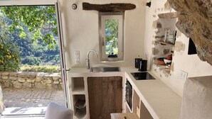 A compact kitchen with a sink, stove, and a small window. There is a chair and a table with a colorful rug. - La Petite Maison (Montaulieu)
