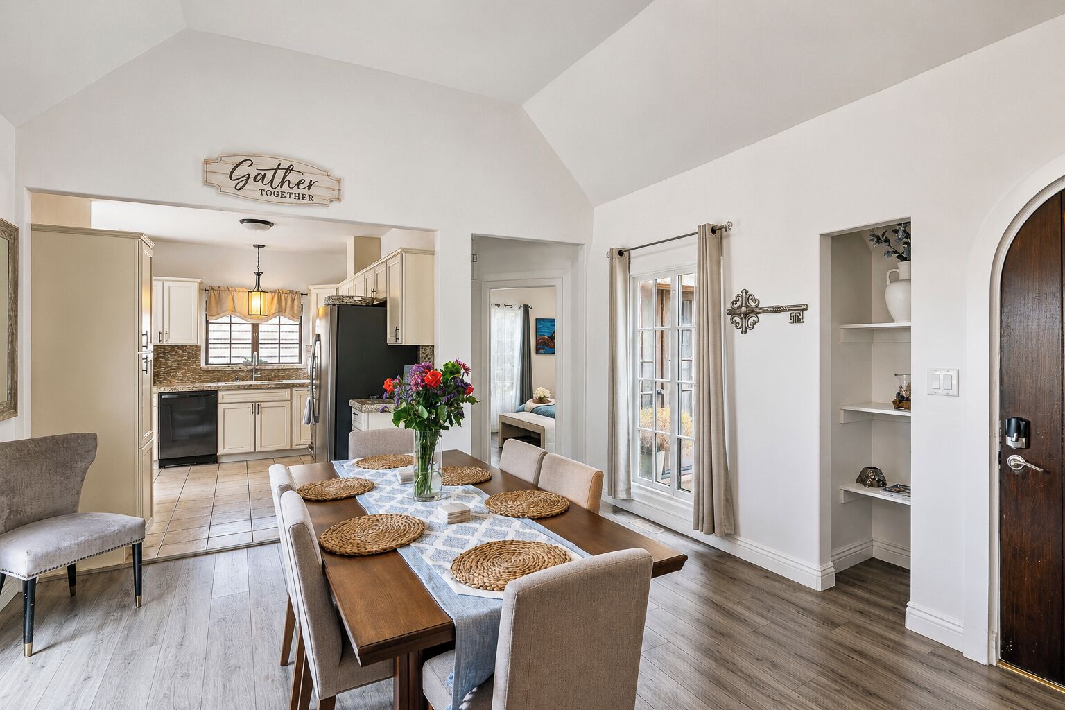 A dining area with a wooden table set for four, a vase with flowers, and a kitchen in the background.