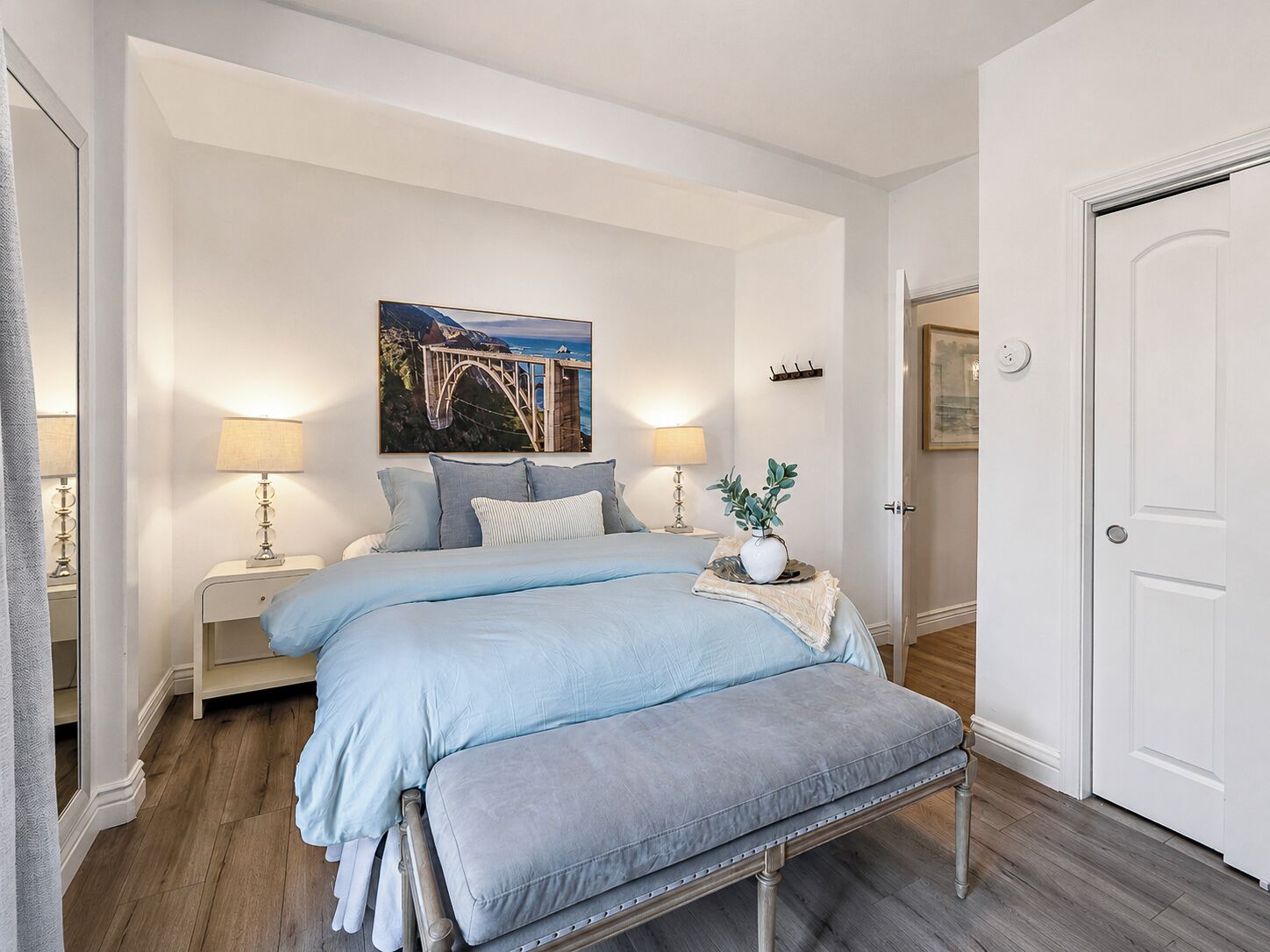 A bedroom with a bed, bedside tables, a bench, a painting, and a door.