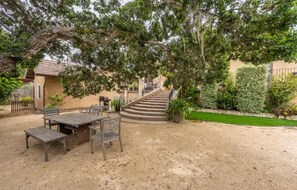A patio with a wooden table and chairs, surrounded by sand and greenery, leading to a staircase. - Large deck, open floor plan, walk to shopping. (Carmel By the Sea)
