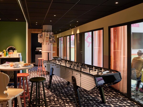 A room with a foosball table, a bar area with stools, and a breakfast setup with various drinks and food items.