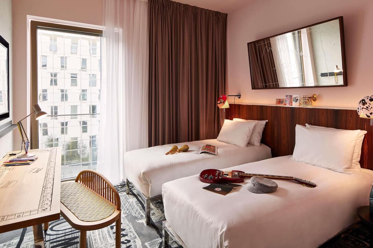 A hotel room with two beds, a desk, a chair, a guitar, and a mirror.