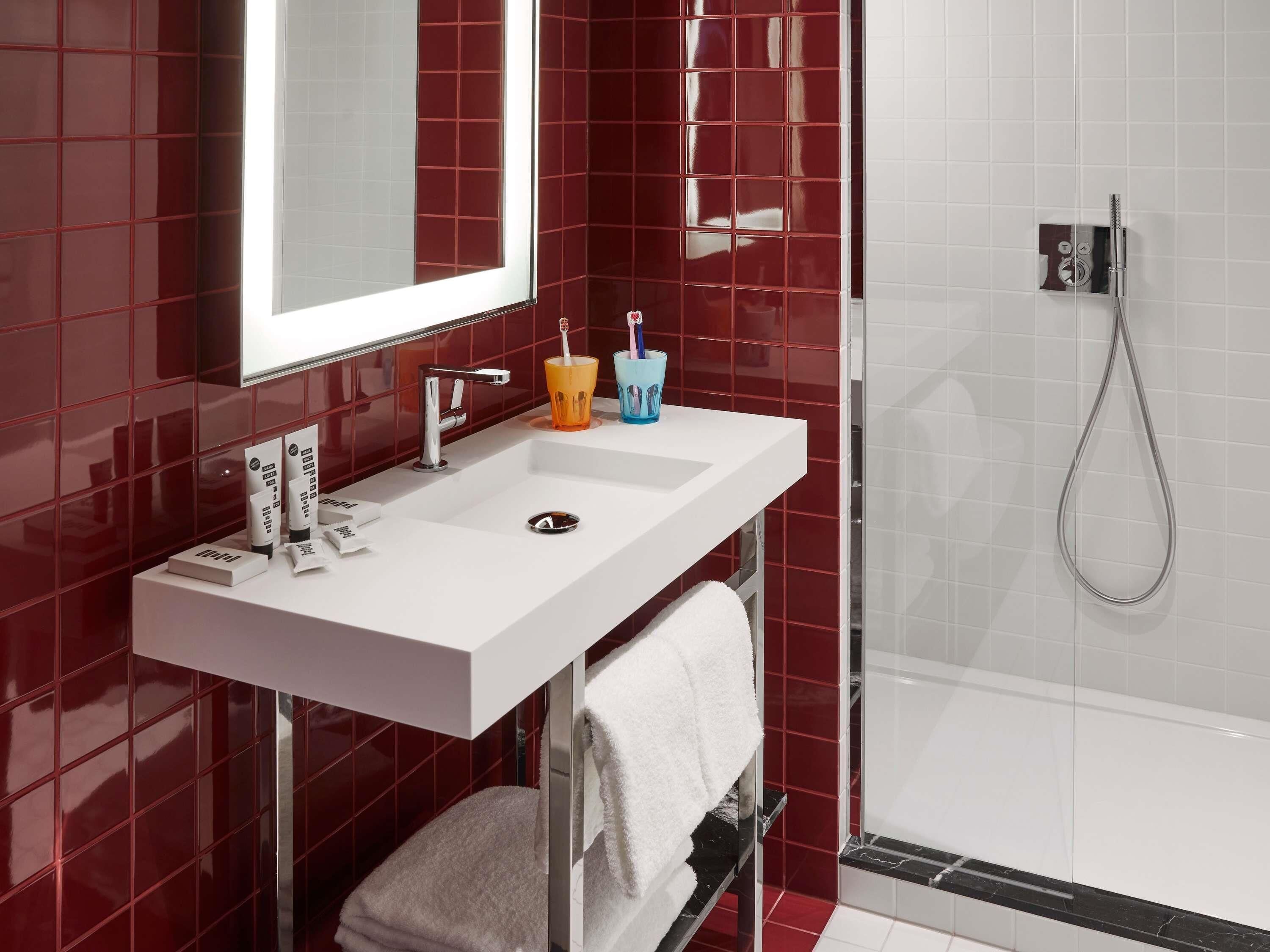 Un baño moderno con lavamanos blanco, paredes de azulejos rojos, cabina de ducha de vidrio y un perchero para toallas.
