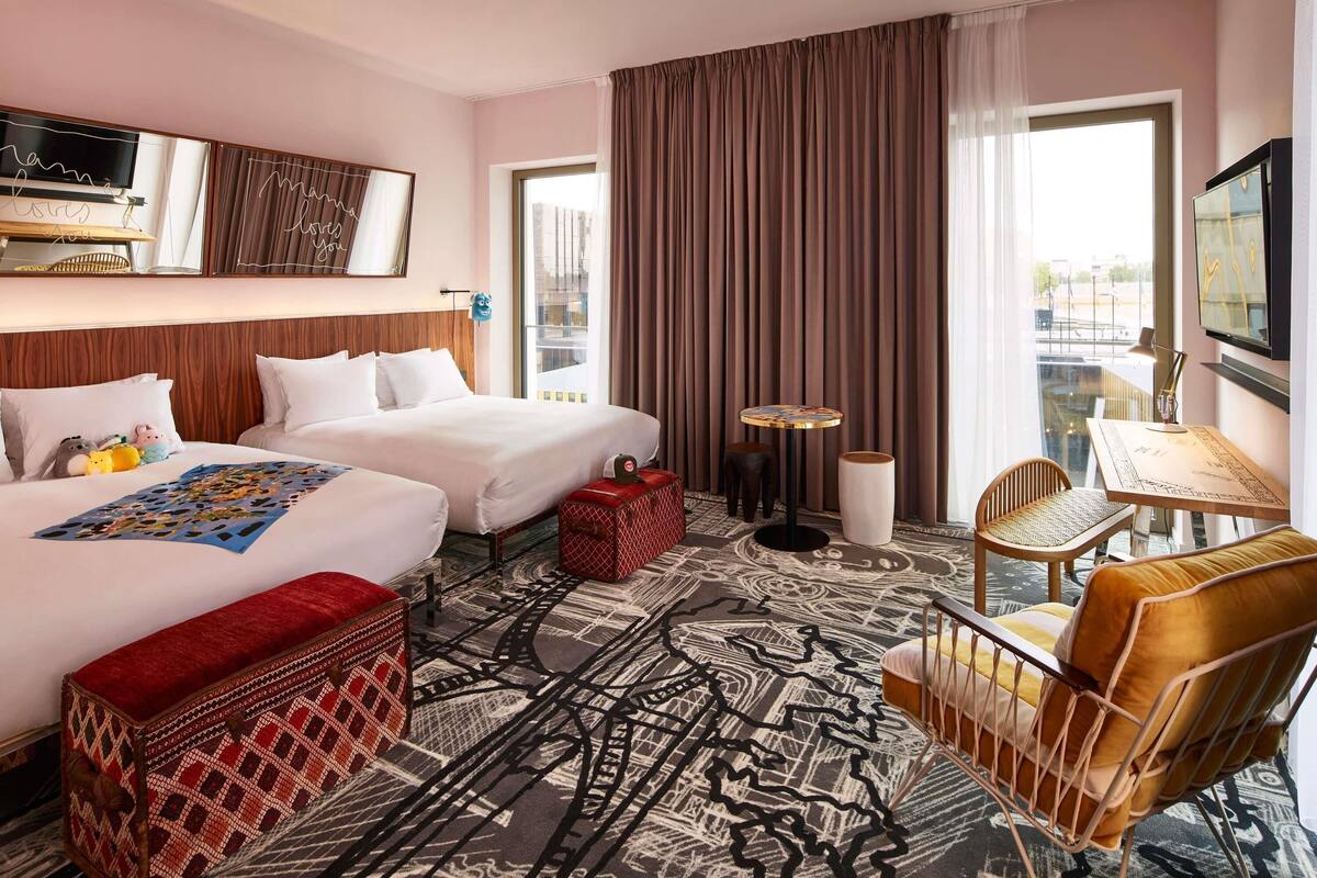 A hotel room with two beds, a small table, a chair, a red ottoman, and a large window with curtains.