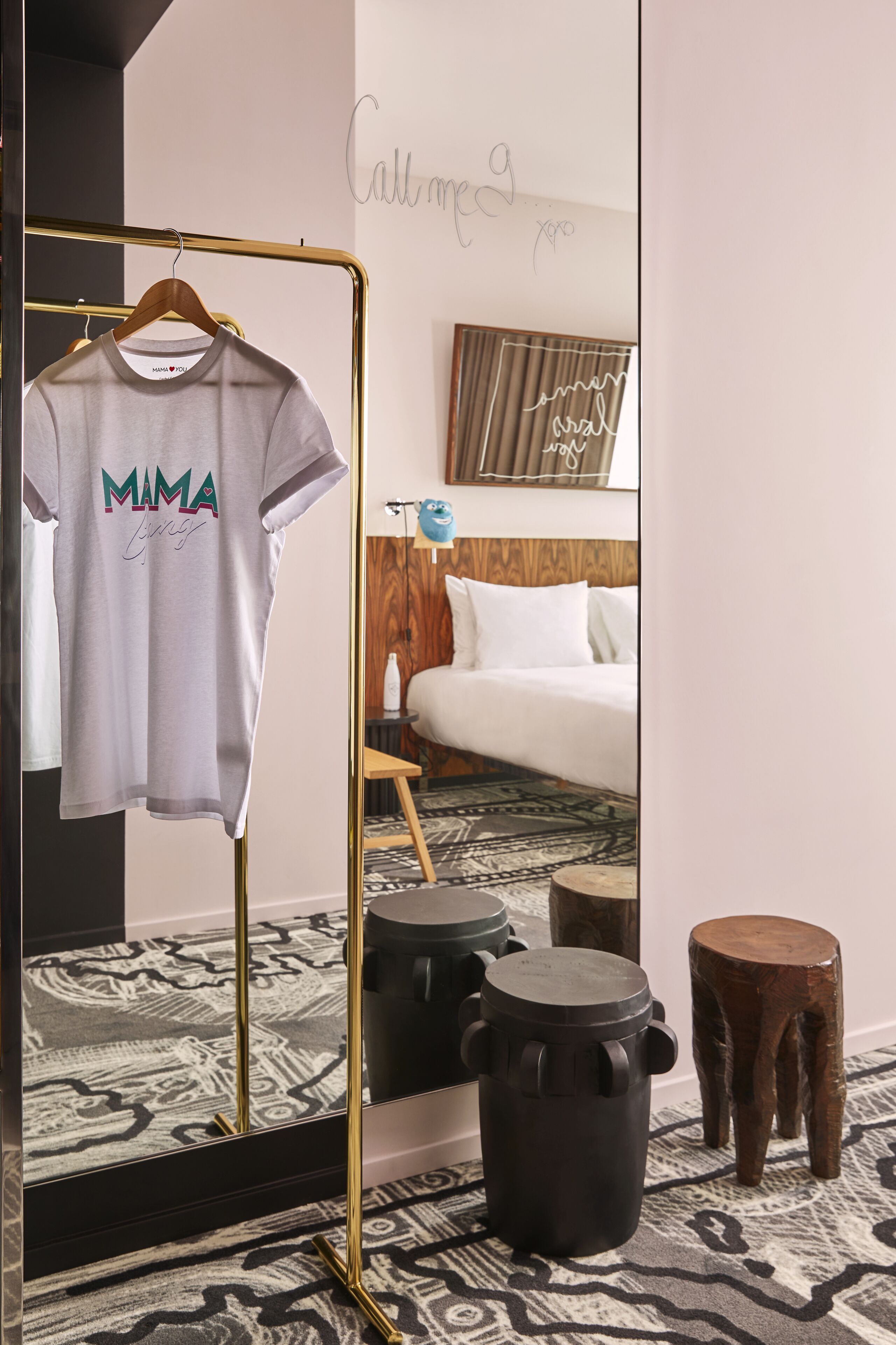 Una habitación de hotel con cama, espejo, perchero con una camiseta, basurero negro y taburete de madera.