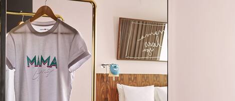 A hotel room with a bed, a mirror, a hanging rack with a t-shirt, a black trash bin, and a wooden stool.