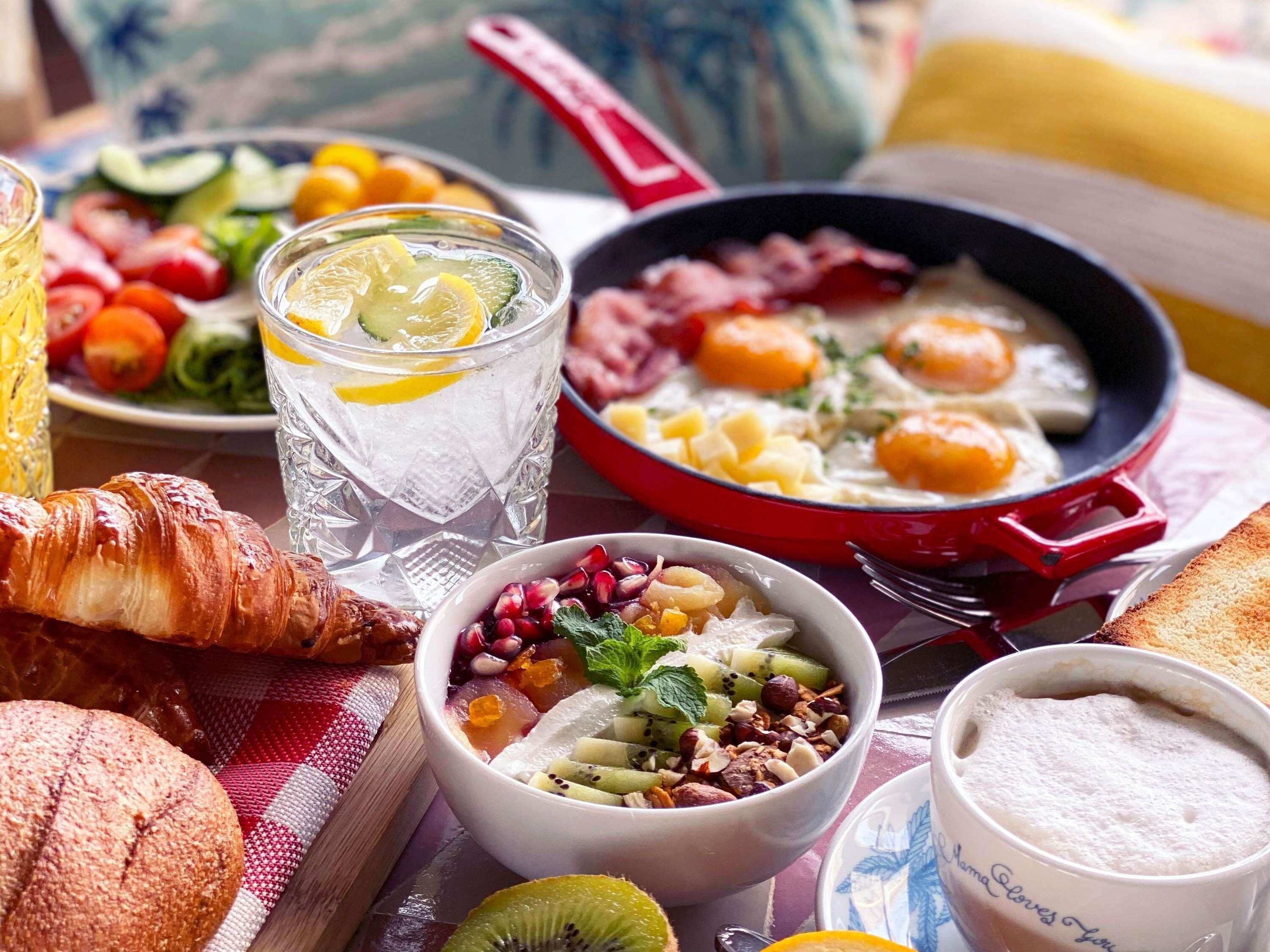 Une table mise pour le petit-déjeuner avec des croissants, un saladier de fruits frais, un verre d’eau glacée, une poêle contenant des œufs et une tasse de café.