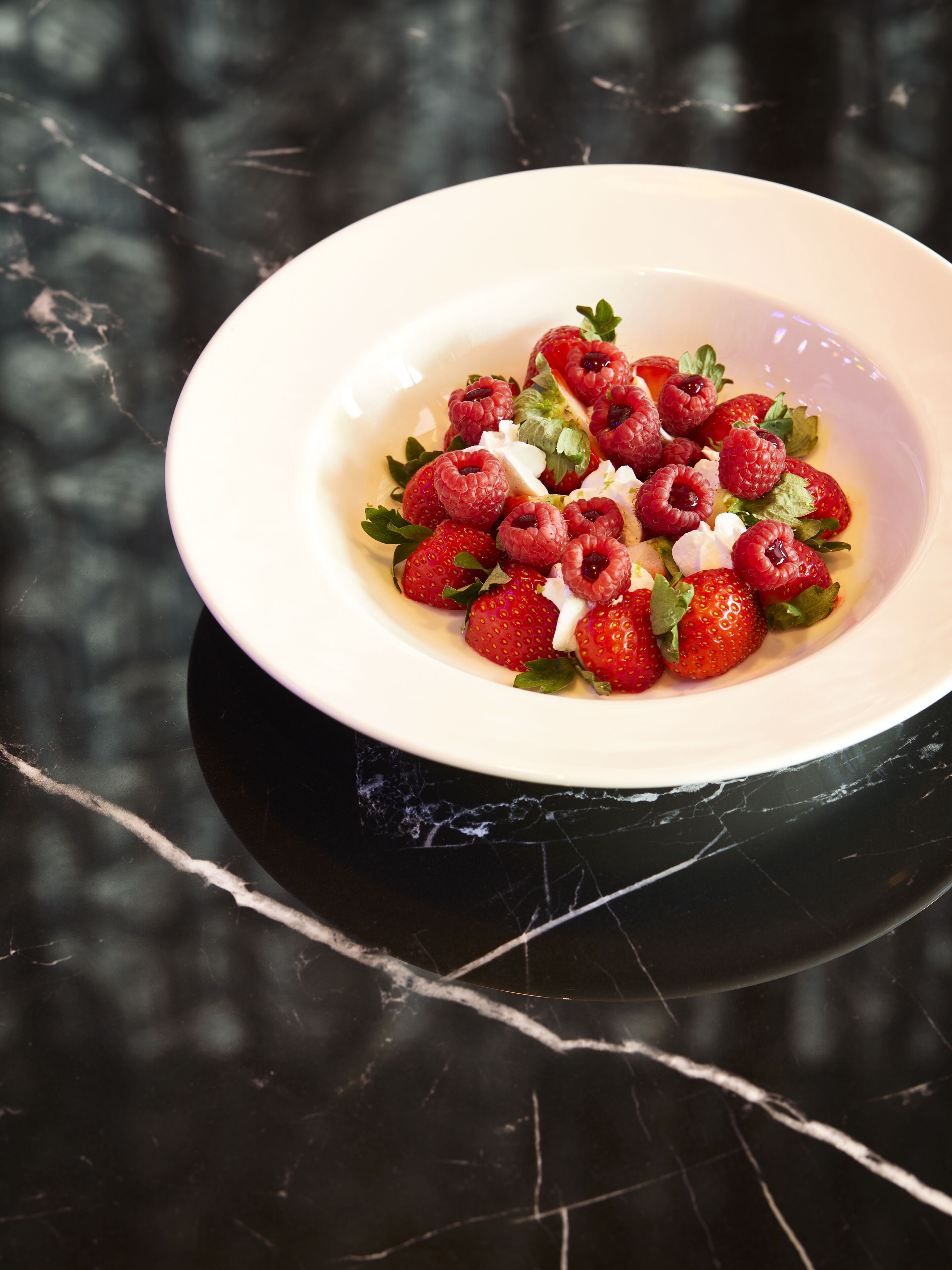 A white bowl with a salad of strawberries and raspberries on a black marble surface.