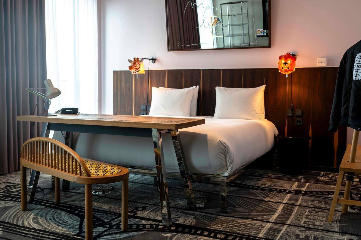 A hotel room with a wooden desk, a chair, a bed with white linens, two wall-mounted lamps, and a large mirror reflecting the room.