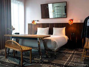 Hotellihuone, jossa on puinen työpöytä, tuoli, valkoisilla lakanoiden peitetty sänky, kaksi seinälle asennettua lamppua ja suuri peili, joka heijastaa huonetta.