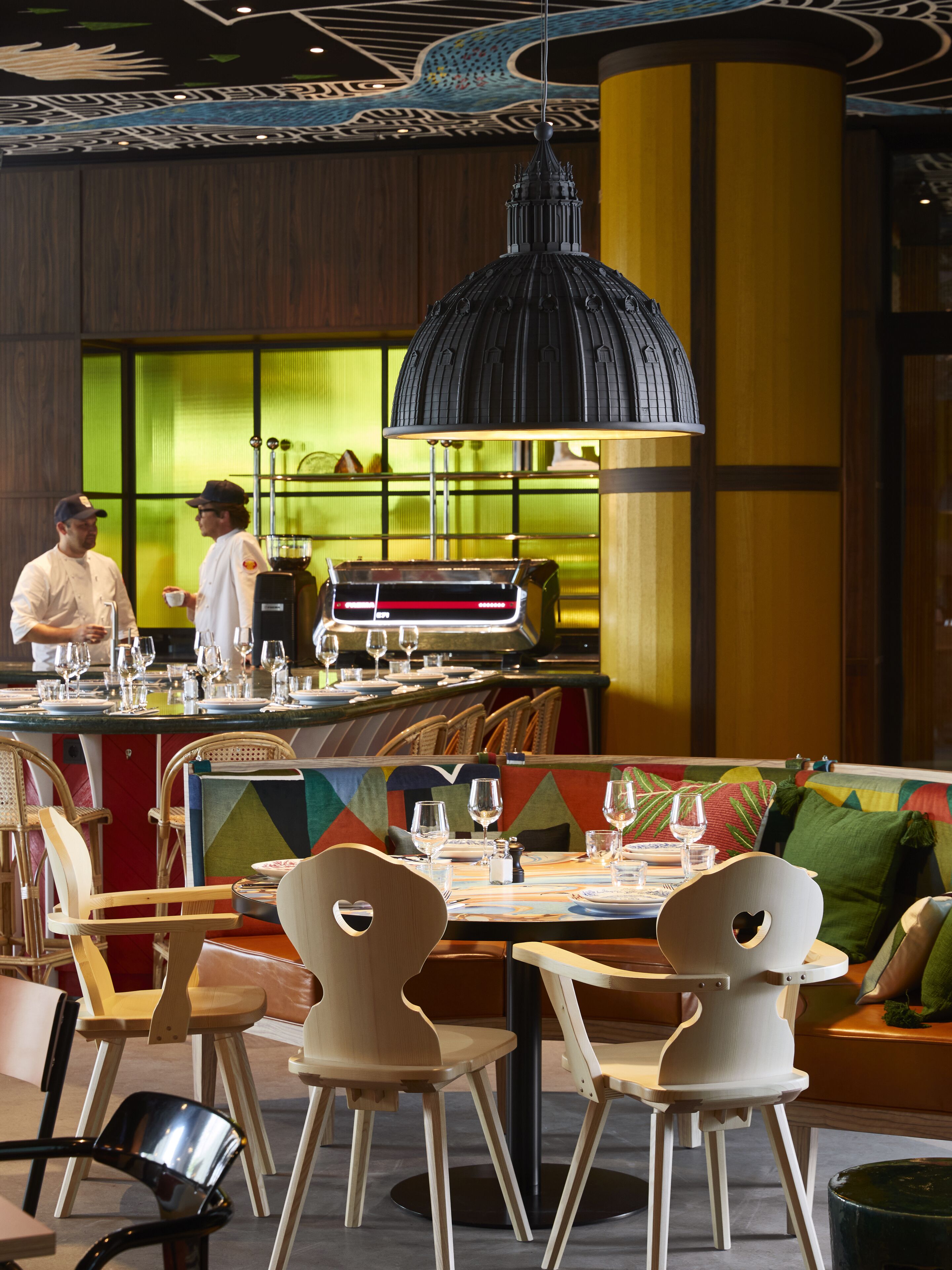 Un restaurante con una decoración moderna, que presenta un área de comedor central con una lámpara colgante negra, sillas de madera y un diseño de pared colorido.
