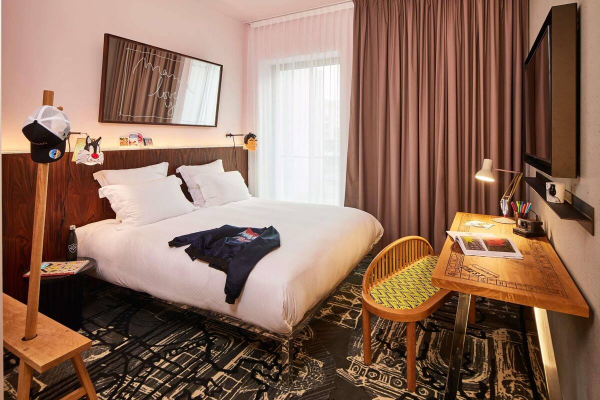 A hotel room with a bed, a desk with a chair, a lamp, a wall-mounted TV, and a window with curtains.