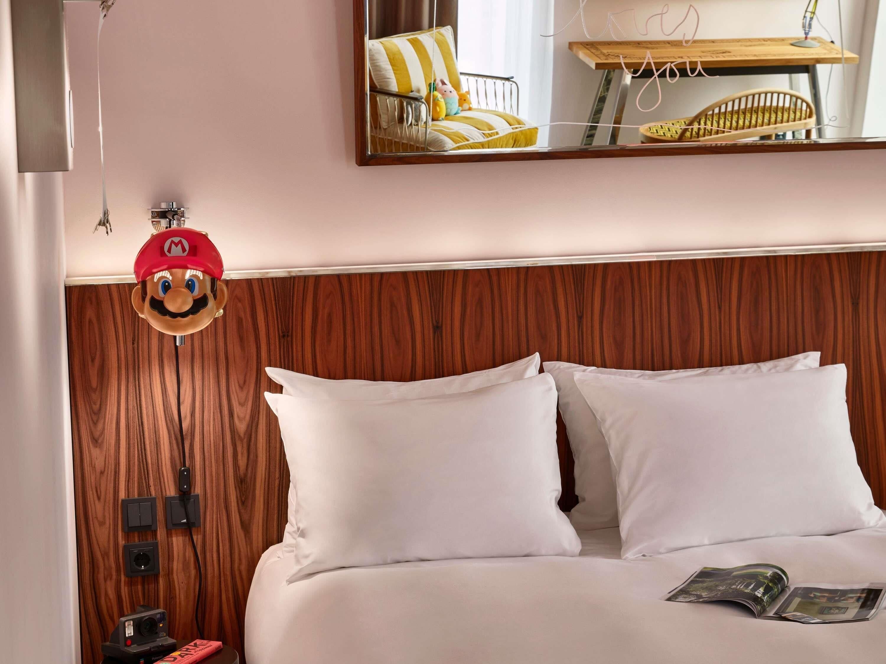 Una habitació d'hotel amb un capçal de llit de fusta, roba de llit blanca, un llit amb una revista i una decoració del personatge Mario.