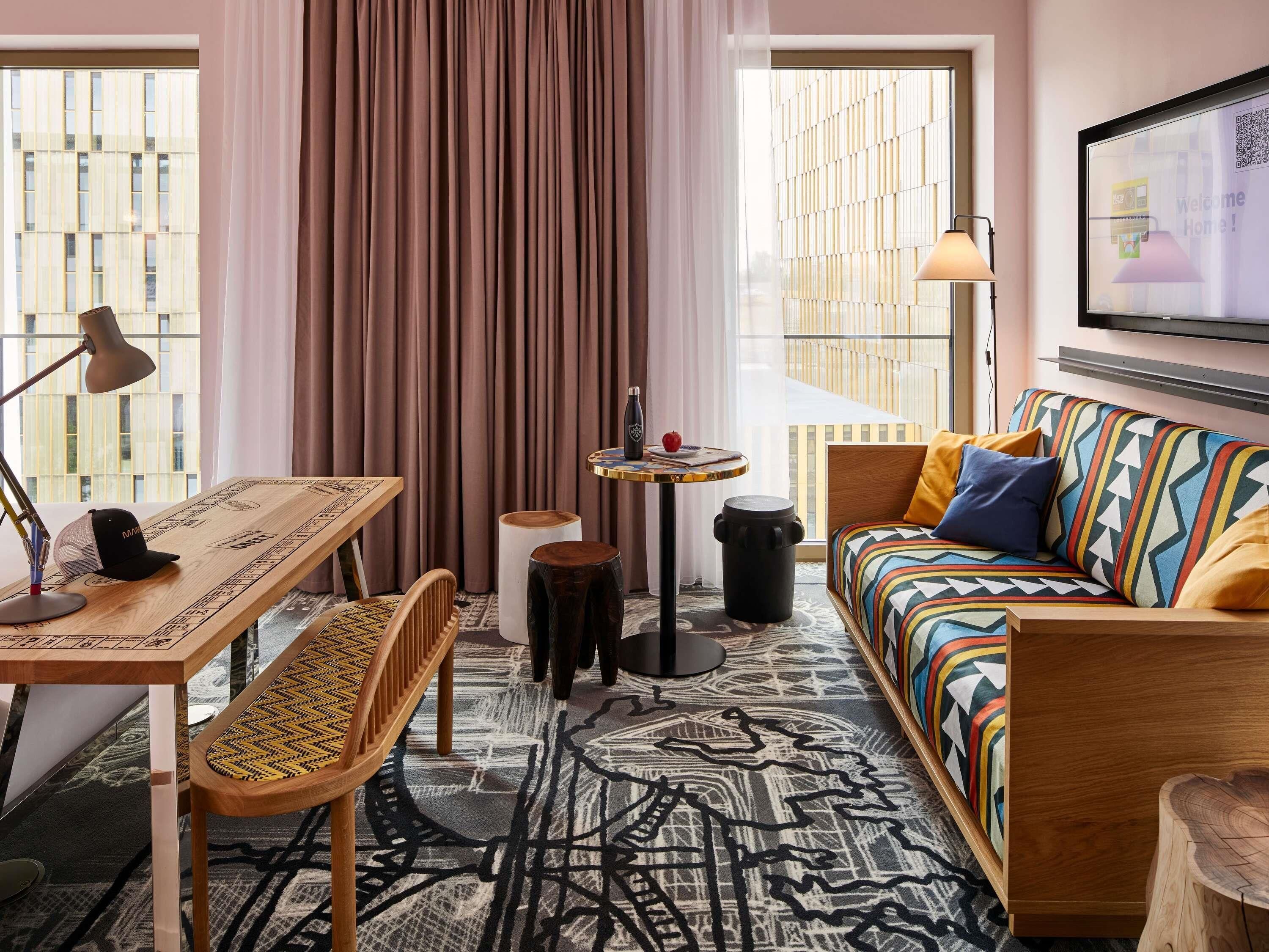 Una habitación de hotel moderna con un escritorio de madera, un sofá colorido, una mesita con una lámpara y un ventanal con cortinas.