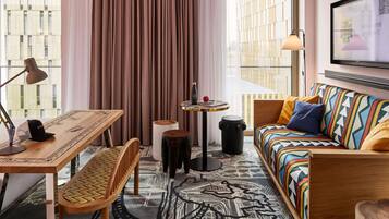 Una habitación de hotel moderna con un escritorio de madera, un sofá colorido, una mesita con una lámpara y un ventanal con cortinas.